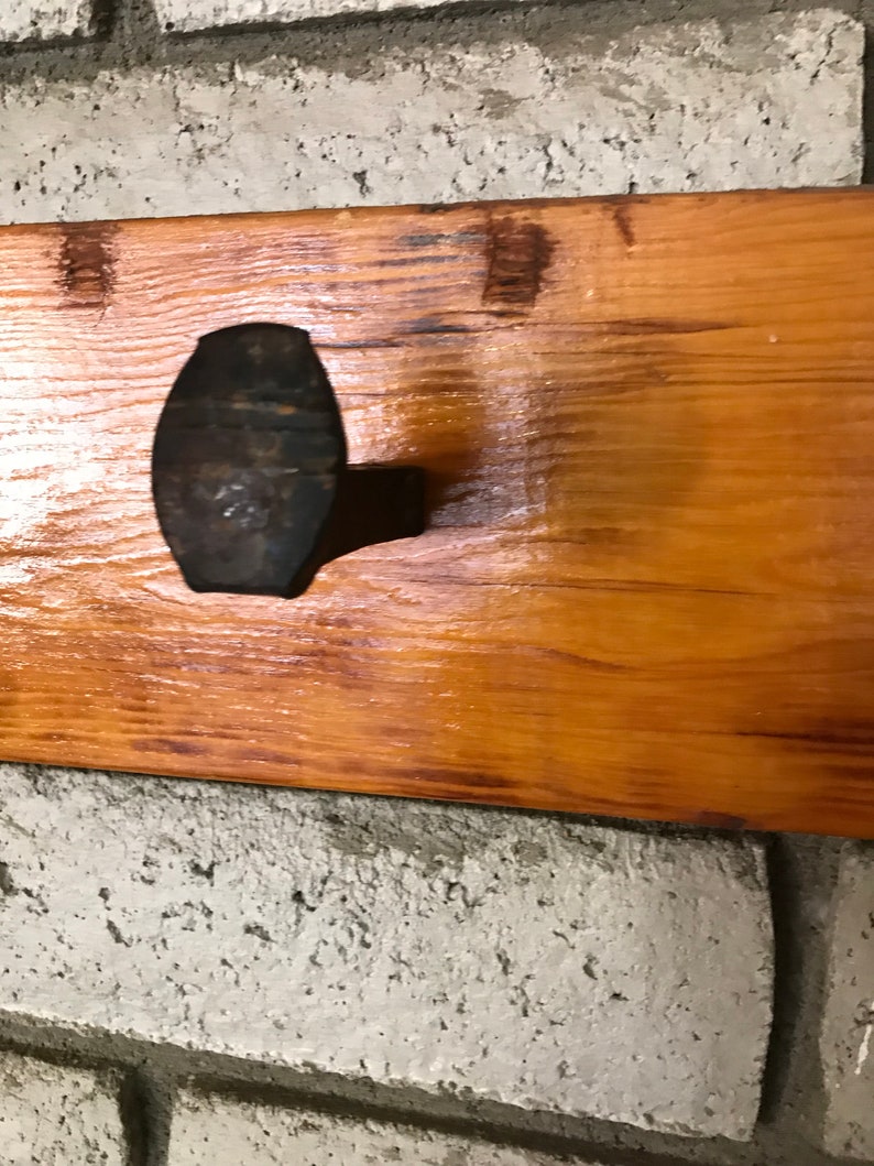 Op de afbeelding: Een rustieke houten wandhaak met een enkele, grote, zwarte metalen haak. De haak is bevestigd aan een houten plank die is gemonteerd op een witte bakstenen muur.