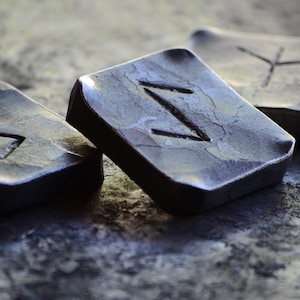 Könnte beinhalten: Drei kleine, rechteckige Metallblöcke mit verschiedenen Symbolen, die in ihre Oberflächen geschnitzt sind. Die Blöcke bestehen aus einem dunklen, metallischen Material und haben eine raue, strukturierte Oberfläche.