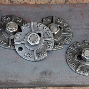 Op de afbeelding: Vier zilveren metalen pijpflensen met een zeskantige moer in het midden van elk. De flensen zijn gemarkeerd met cijfers en letters, waaronder "1/2", "NO", "MA", "15 & 10", "30" en "6810".