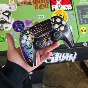 May include: A black and white video game controller with a colorful, pixelated design. The controller has a black base with white buttons and a touchpad. The design features a mix of purple, teal, and orange colors.