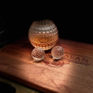 Op de afbeelding: Een houten snijplank met een gepersonaliseerde inscriptie "SEAN October 28th, 2017". Drie doorzichtige glazen voorwerpen in de vorm van een golfbal liggen op de plank, een is een groter glas met een bruine vloeistof erin.