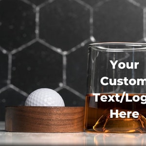 Op de afbeelding: Een helder glas met een bruine vloeistof erin. Het glas heeft de tekst "Your Custom Text/Logo Here" erop gedrukt. Een golfbal ligt op een houten onderzetter.