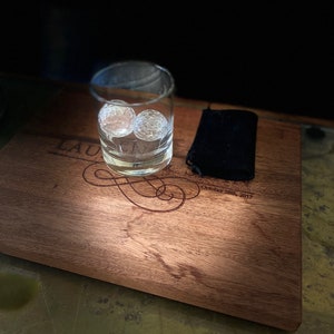 Op de afbeelding: Een houten serveerblad met een gepersonaliseerde inscriptie die "Lauren & Mark October 28th, 2017" luidt. Een glas met twee heldere ijsbollen staat op het blad, naast een zwarte doek.