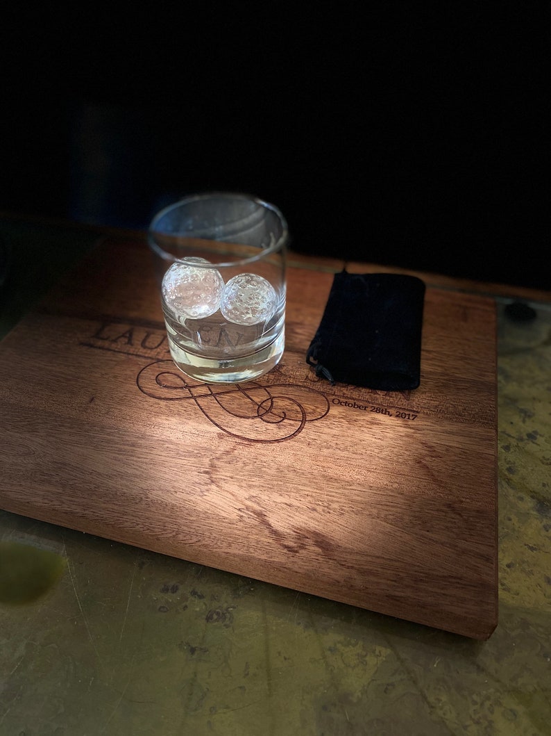 Op de afbeelding: Een houten serveerblad met een gepersonaliseerde inscriptie die "Lauren & Mark October 28th, 2017" luidt. Een glas met twee heldere ijsbollen staat op het blad, naast een zwarte doek.