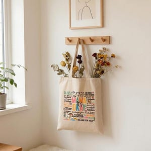 Può includere: Una borsa tote in tela beige con il testo "Mama It's Okay" in vari colori. La borsa è appesa a un gancio da parete in legno, piena di fiori secchi. Un disegno a tratto incorniciato è appeso sopra la borsa.