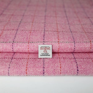 Könnte beinhalten: Ein gefaltetes Stück rosa Harris-Tweed-Stoff mit einem dezenten Karomuster aus dünnen violetten und orangefarbenen Linien. Ein weißes rechteckiges Etikett ist in der Mitte aufgenäht und trägt die Aufschrift Hand Woven Cloth Harris Tweed Outer Hebrides of Scotland.
