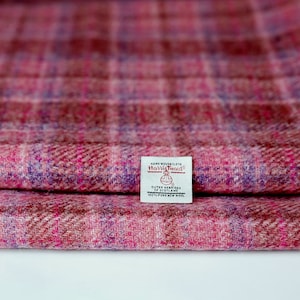 Könnte beinhalten: Ein gefaltetes Stück Harris Tweed Stoff mit rosa, rotem und violettem Karomuster. Ein weißes rechteckiges Etikett ist auf den Stoff genäht und trägt den Text: Hand Woven Cloth, Harris Tweed, Outer Hebrides of Scotland, 100% Pure New Wool.