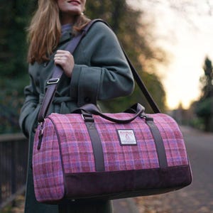 Könnte beinhalten: Eine rosa-lila karierte Reisetasche mit braunen Lederdetails. Die Tasche hat einen Schultergurt und Griffe. Der Boden der Tasche ist einfarbig dunkelbraun. Das Etikett lautet "Harris Tweed".