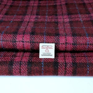 Könnte beinhalten: Ein gefaltetes Stück Harris Tweed Stoff mit rotem, schwarzem und blauem Karomuster. Ein weißes rechteckiges Etikett ist in der Mitte aufgenäht und trägt den Text: Hand Woven Cloth, Harris Tweed, Outer Hebrides of Scotland, 100% Pure New Wool.