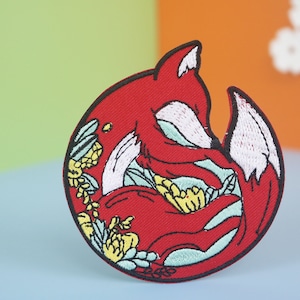 May include: Red embroidered patch of a curled up fox with floral details. The fox is surrounded by green and yellow flowers.