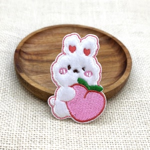 May include: A white bunny patch with red heart accents on its ears and cheeks, holding a pink peach with a green leaf. The patch has a red stitched outline and is set on a wooden plate.
