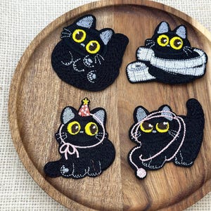 May include: Four embroidered black cat patches with large yellow eyes. One cat is curled up, one is wrapped in a white measuring tape, one wears a colorful party hat with a pink ribbon, and one plays with a pink ball of yarn and string.