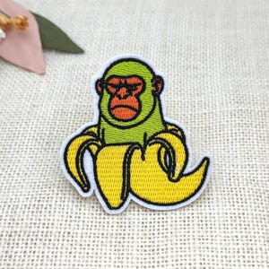 May include: Embroidered patch featuring a green monkey with an orange face and a yellow banana body. The patch has a white border and is set against a textured, neutral background. Decorative floral elements are visible in the upper left corner.