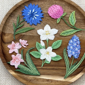 Pode incluir: Uma bandeja de madeira exibe vários patches florais bordados. Os patches incluem uma flor azul, um trevo rosa, flores brancas e um jacinto azul. Folhas e caules verdes acompanham as flores, criando um arranjo artesanal colorido.