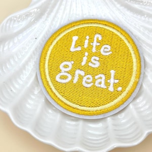 Puede incluir: Un parche bordado amarillo con las palabras "Life is Great" en letras blancas. El parche es redondo y tiene un borde blanco. Se coloca sobre un plato blanco con volantes.
