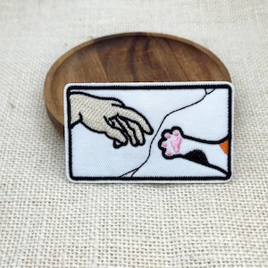 May include: A rectangular embroidered patch featuring a white background with a black border. The design depicts a human hand reaching out to touch a cat paw, with the cat paw showing pink pads and black and orange fur markings on a white base.