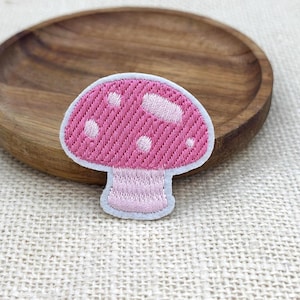 Può includere: Una toppa ricamata a forma di fungo con un cappello rosa a pois bianchi e un gambo rosa chiaro. La toppa presenta un bordo bianco ed è realizzata con un motivo a filo testurizzato, creando una silhouette di fungo ben definita.