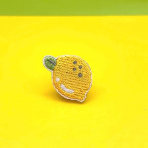 May include: Embroidered lemon patch with a yellow body, small green leaf, and white outline. The lemon has three small grey dots and a white smile. The patch is on a bright yellow background with a green top.