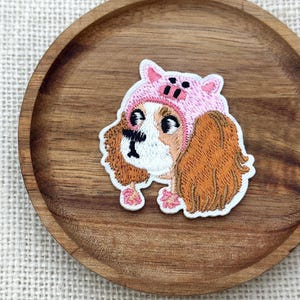 May include: An embroidered patch featuring a brown and white spaniel dog wearing a pink pig hat. The dog has long, textured brown ears, black eyes, and a white muzzle. The pig hat is light pink with small ears and a snout detail on the forehead.