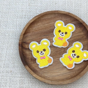 May include: Three yellow bear patches with light blue borders and pink bow ties are arranged on a wooden plate. The patches have black eyes and noses. The plate is on a textured, off-white surface.