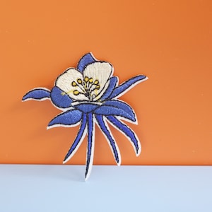 May include: An embroidered flower patch in blue and white with a white background. The flower has five petals and a yellow centre.