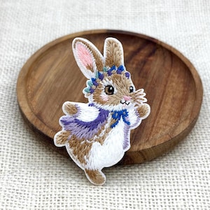May include: An embroidered iron-on patch featuring a brown and white rabbit with a pink inner ear, wearing a purple and blue floral crown and a matching purple and blue cape. The rabbit is depicted in a dynamic pose with its arms outstretched.