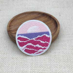 Puede incluir: Un parche bordado circular con un diseño de paisaje. La sección superior muestra un cielo rosa pálido con un sol blanco, una banda central morada y una sección inferior de colinas onduladas de color rosa oscuro con reflejos blancos, bordeado en blanco.