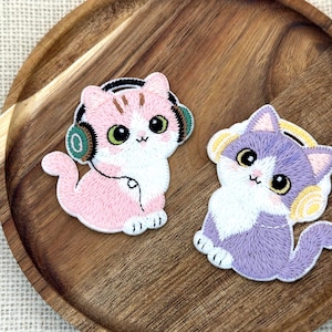 May include: Two embroidered cat patches. One is pink and white with green headphones, the other is purple and white with yellow headphones. Both have large eyes and are on a wooden tray.