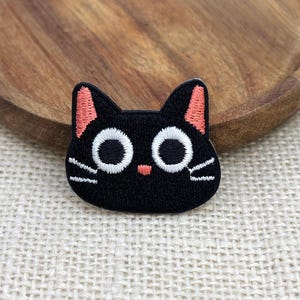 May include: An embroidered patch shaped like a black cat head with large white circular eyes, a small pink nose, pink inner ears, and white stitched whiskers on each side. The patch features a dense black thread texture throughout the design.