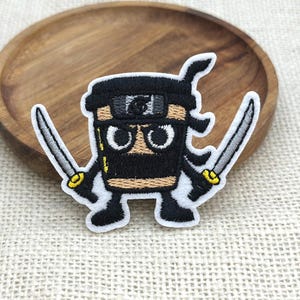 Pode incluir: Um emblema bordado com uma personagem de desenho animado que se assemelha a um copo de café vestido de ninja. A personagem é preta e bege com olhos brancos grandes, uma faixa com um símbolo e duas espadas prateadas nas mãos.