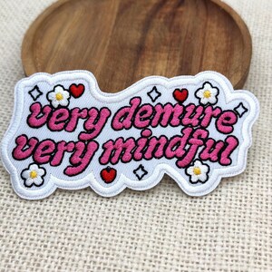 May include: Embroidered patch with the words "very demure very mindful" in pink script. The patch is white with a scalloped edge and features small flower and heart designs. The patch is on a textured surface.