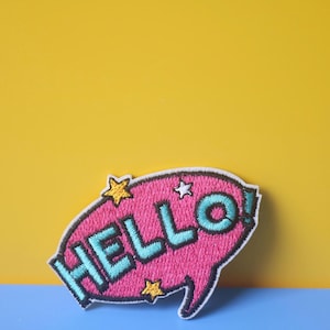 May include: Pink and blue embroidered patch with the word "HELLO!" in a speech bubble with yellow stars.