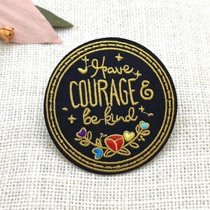 May include: A circular black embroidered patch featuring gold stitched text that reads I have COURAGE & be kind. The design includes a gold double-stitched border, small gold stars, and a floral motif at the bottom with red, yellow, and blue accents.