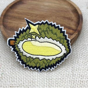 Könnte beinhalten: Ein gestickter Aufnäher, der eine stilisierte Durianfrucht mit einer stacheligen olivgrünen Außenseite, einem weißen Rand und einem hellgelben Innenteil zeigt. Über der Frucht befindet sich ein kleiner gelber Stern und links ist ein brauner Stiel zu sehen.