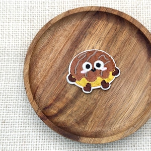 May include: A brown and yellow embroidered patch in the shape of a cartoon character with large eyes and small feet. The patch is on a round wooden plate with a light-colored woven background.