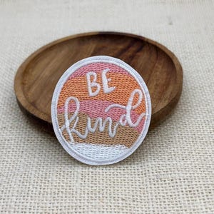 May include: A circular embroidered patch featuring horizontal stripes in shades of orange, pink, and tan. The text "BE KIND" is stitched in white thread across the center, with "BE" in a block font and "KIND" in a cursive script font.