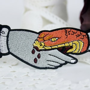 May include: Embroidered iron-on patch featuring a snake biting a hand with blood dripping from the wound. The snake is orange and yellow, and the hand is gray.