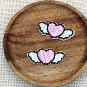 May include: Two embroidered patches, each featuring a light pink heart with white wings. The hearts have a subtle sparkling effect and are outlined in dark brown thread. The wings are white with dark brown edges, creating a cohesive design.