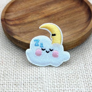 May include: An embroidered patch featuring a light blue cloud with a smiling face, pink cheeks, and blue Zz text. A yellow crescent moon with a closed eye rests on top of the cloud. The design is stitched with white, blue, yellow, and pink thread.