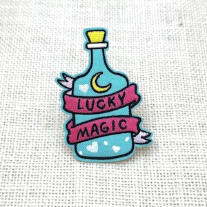 Puede incluir: Un parche turquesa con forma de botella con un corcho amarillo y una pancarta rosa que dice "LUCKY MAGIC". La botella tiene una luna creciente y corazones blancos. El parche está delineado en negro.