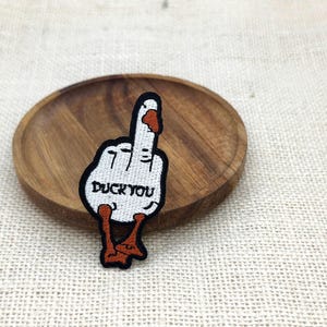 Pode incluir: Um emblema bordado apresentando um ganso branco com bico e patas laranjas, moldado como uma mão fazendo um gesto de dedo médio. O ganso tem contornos pretos e o texto DUCK YOU bordado em preto no centro do seu corpo.