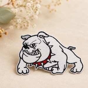 Peut inclure: Un écusson brodé représentant un bouledogue blanc à l'expression féroce, avec des contours noirs et un collier rouge à pointes. Le motif montre le chien dans une posture basse et agressive avec des dents visibles, réalisé en fil détaillé.