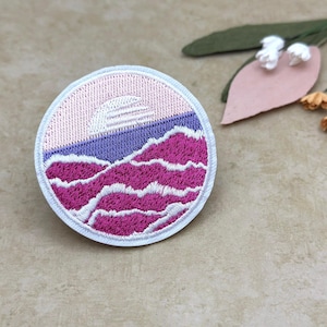 Puede incluir: Parche bordado con un diseño de paisaje. El parche presenta un cielo rosa, un horizonte púrpura y azul, y montañas magenta. El parche es redondo con un borde blanco. El diseño recuerda una puesta de sol sobre montañas.