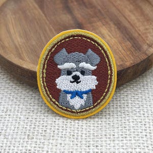 May include: Embroidered patch featuring a Schnauzer dog with gray fur, a white beard, and a blue bow tie. The patch has a brown oval center with a gold border, set against a yellow outer edge. The patch is likely for clothing or accessories.