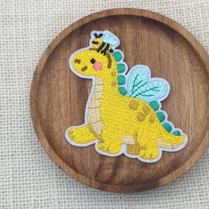 May include: Embroidered patch featuring a yellow dinosaur with a bee on its head and blue wings. The dinosaur has green spines and brown spots. The patch is on a wooden plate.