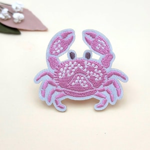 May include: Embroidered patch of a pink crab with a light blue border. The crab has detailed stitching on its body and claws. This patch is suitable for decorating clothing, bags, or other fabric items.