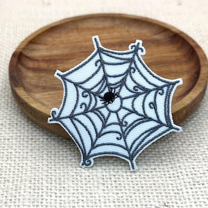 Puede incluir: Un parche bordado que presenta un diseño de telaraña azul claro con costuras gris oscuro y una pequeña araña negra en el centro. El parche tiene una forma hexagonal distintiva con bordes decorativos rizados en cada uno de los seis puntos.