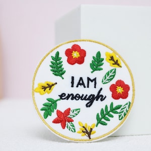 Op de afbeelding: Witte geborduurde patch met een gouden rand. De patch heeft een bloemenmotief met rode, gele en groene bloemen en bladeren. De tekst "I AM enough" is in zwart geborduurd.