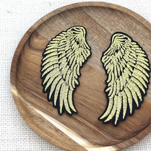 May include: A pair of embroidered patches featuring detailed golden yellow wings with black outlines. The wings are shaped with individual feather stitching patterns, creating a textured appearance. These two separate wing-shaped patches are designed for application onto fabric surfaces.