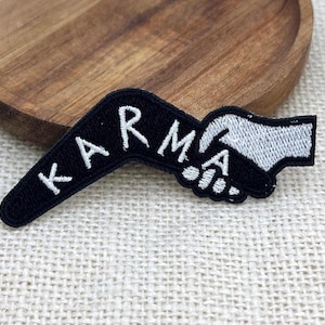 May include: A black embroidered patch featuring the word KARMA in white capital letters, followed by a graphic of two hands shaking. The patch has a textured fabric surface and a distinct border, showcasing a high-contrast design in black and white.